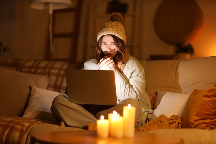 Frozen young woman in warm clothes using laptop at home during b